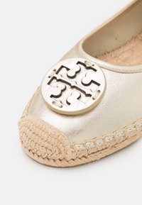 Bailarina metálica dorada con acento de logotipo redondo y recortado, y una puntera de yute tejido beige con detalles de costura.