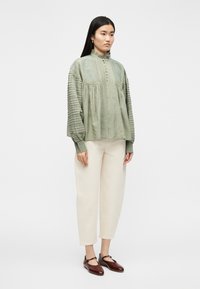 Γυν αίκα που φοράει μπλούζα σε πράσινο φασκόμηλο με υφή και κουμπιά, ανοιχτό μπεζ cropped παντελόνι και σκούρα καφέ παπούτσια Mary Jane, στέκεται μπροστά σε λευκό φόντο.