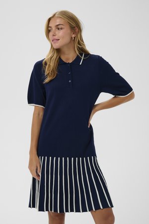 Vestido polo de color navy con mangas cortas, frente abotonado y falda plisada a rayas blancas. Textura de punto suave con detalle de cuello en contraste.