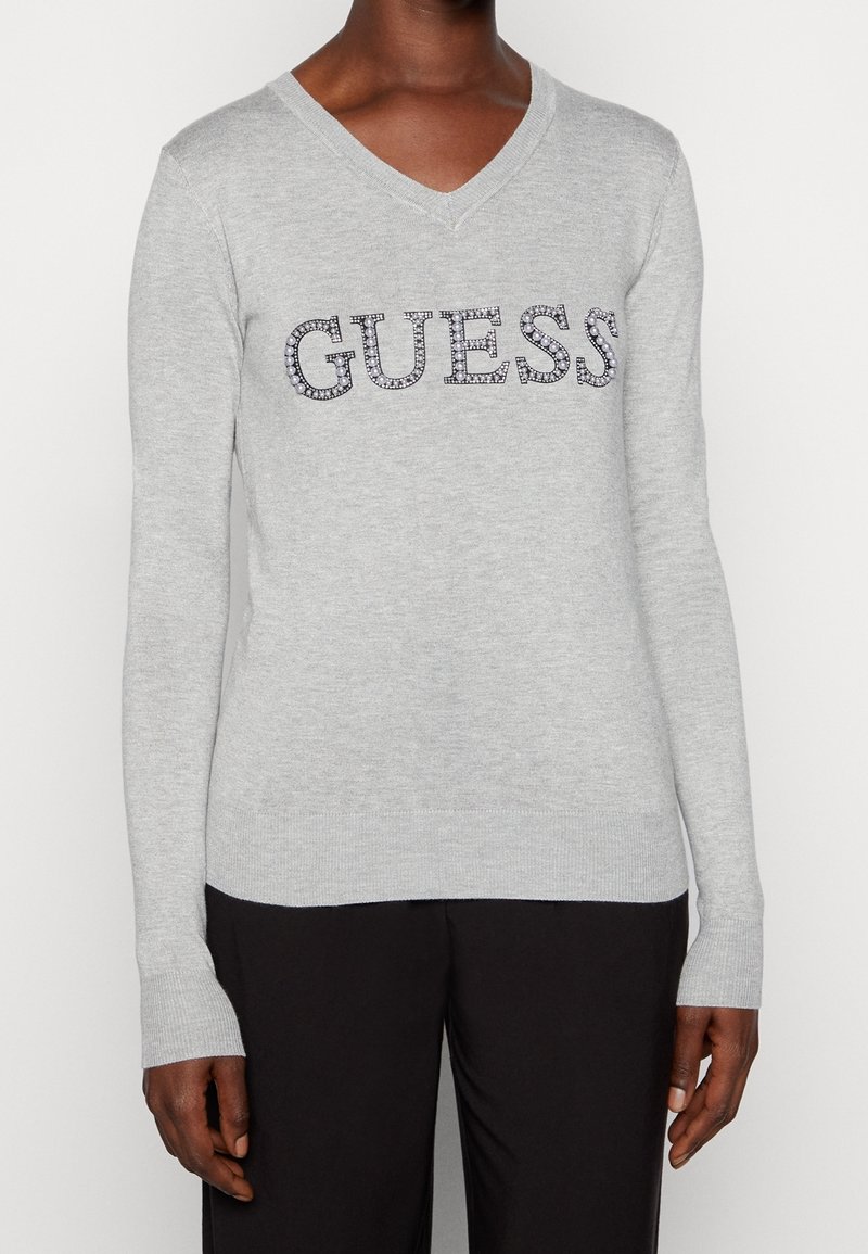 Personne portant un pull gris clair à col en V avec "GUESS" en strass sur la poitrine et un pantalon noir, debout devant un fond blanc.