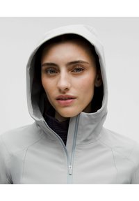 Chaqueta con capucha de color gris claro con cremallera, que presenta un cuello alto y una textura suave. La capa interior es de color púrpura oscuro con detalles acolchados.