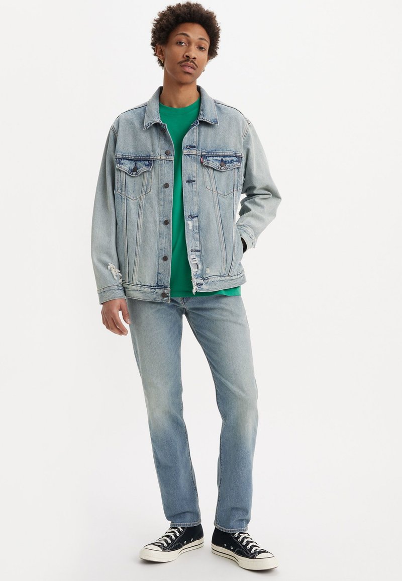 Giacca di jeans azzurro chiaro con due tasche sul petto, abbinata a una T-shirt verde e jeans chiari. Sneakers nere con suole bianche.