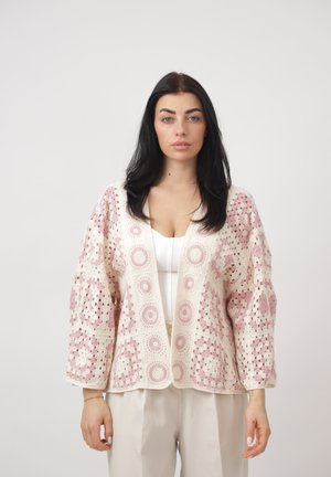 Kvinde med langt mørkt hår iført en hvid top og beige bukser, iført en creme- og lyserød hækleet cardigan med åben front.