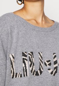 Szary sweter z dzianiny z napisem "LIU" w zwierzęcy wzór z cekinami. Szeroki dekolt i miękka faktura. Zbliżenie na szczegóły wzoru.