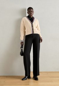 Cardigan cremoso a maglia con bordi a strisce colorate, abbinato a pantaloni neri a vita alta e stivaletti neri. Inclusa piccola borsa nera.