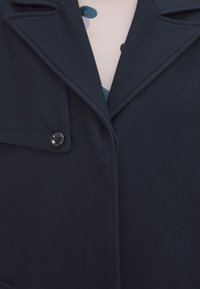 Chaqueta azul marino fabricada en tejido texturizado con cuello de solapa, que cuenta con un solo botón negro y detalles de costura visibles.