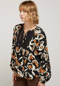 Blouse noire avec un imprimé animal abstrait en orange et crème, dotée de manches longues, d'un col rond et d'un détail à lacets à l'avant.