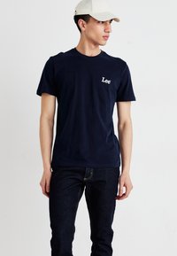 Marineblå bomull t-skjorte med korte ermer og rund hals, med en hvit "Lee" logo på venstre bryst. Brukt sammen med mørke jeans.