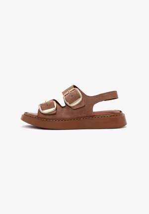 Sandale en daim marron avec des sangles à double boucle réglables, talon découvert et semelle épaisse marron. Design plat et minimaliste avec des détails cousus.