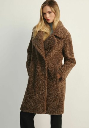 Kobieta z długimi blond włosami, ubrana w brązowy, teksturowany płaszcz teddy z dużym kołnierzem, z rękami w kieszeniach, do tego czarne spodnie i szary golf.