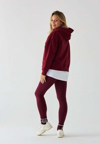 Bordowa bluza z kapturem z kieszenią na przodzie, noszona na białej koszuli; połączona z fakturowanymi bordowymi legginsami i białymi butami sportowymi.