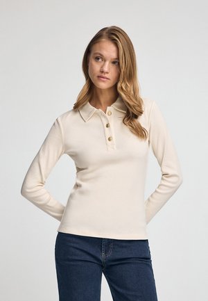 Beige long-sleeve top with a collared neckline, three gold buttons, and a fitted design. Paired with dark blue jeans.