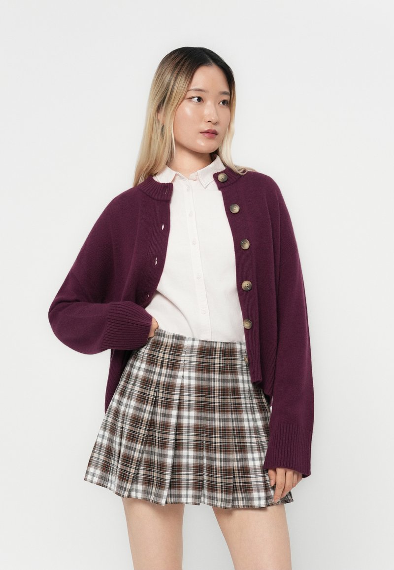 Fioletowy kardigan robiony na drutach z dużymi guzikami, noszony na jasnej koszuli i mini spódnicy w kratę z brązowymi i białymi wzorami. Gładka struktura.