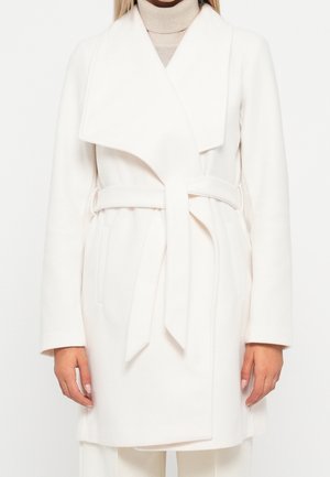 Kvinde iført hvidt bindebælte frakke over en beige højhalset sweater, stående mod en ensfarvet hvid baggrund.