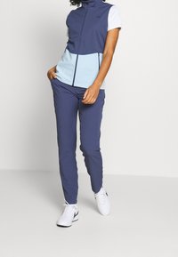 Navy and light blue zip-up vest with high collar, paired with navy pants and white athletic shoes. Fabric appears smooth and fitted.