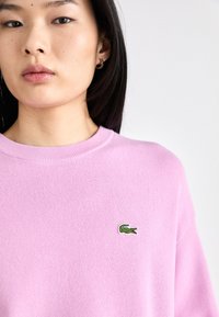 Rózsaszín kötött pulóver, kerek nyakkal és rövid ujjakkal, bal oldalon egy kis zöld krokodil logóval hímezve.