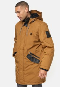 Parka marrón con capucha, fabricada en un tejido texturizado, cierre frontal de cremallera y detalles de bolsillo tipo parche en negro. Incluye cordones y un ajuste cómodo.
