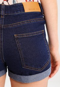 Pantalones cortos de mezclilla con un lavado azul oscuro, con puños doblados y costuras claras. Una etiqueta de cuero marrón en la parte trasera de la cintura dice "Noisy May".