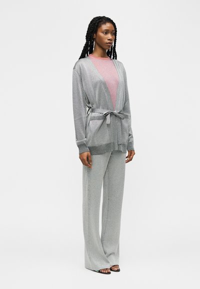 Γυναίκα με μακριά κοτσίδα, φοράει ανοιχτό γκρι ζακέτα με ζώνη, παντελόνι φαρδύ στο ίδιο χρώμα, ροζ μπλούζα και μαύρα σανδάλια με ανοιχτά δάχτυλα, στέκεται πλαγίως.