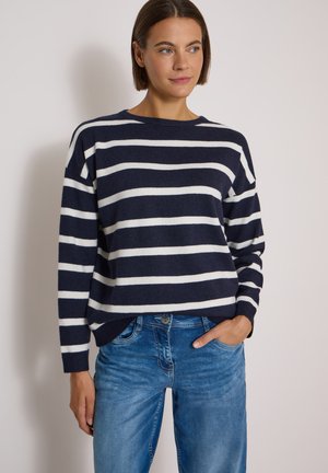Femme portant un pull rayé bleu marine et blanc à manches longues et un jean bleu, debout, une main dans la poche, contre un mur uni.