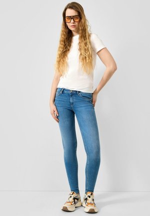Frau mit langen welligen Haaren, die orange getönte Brille, weißes T-Shirt, blaue Skinny Jeans und mehrfarbige Sneakers trägt, steht vor einem einfarbigen Hintergrund.