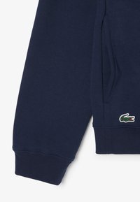 Felpa blu navy con polsini e orlo a costine, caratterizzata da un piccolo logo di coccodrillo verde e rosso sul lato sinistro. Tessuto morbido e texture.