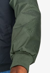 Zelená vodoodpudivá tkanina s texturovaným povrchem a elastickými ribovými manžetami. Detail kapek na rukávu zvyšuje funkčnost.