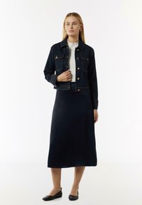 Dunkelblaue, taillierte Jeansjacke mit goldenen Knöpfen, getragen über einem hellen Oberteil und einem langen, dunklen, glatten Rock. Schwarze flache Schuhe runden den Look ab.