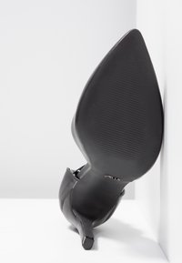 Chaussure à talon haut noire à bout pointu, inclinée avec la semelle tournée vers l'avant sur un fond blanc.