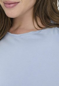 Lyseblå, myk stofftopp med vid hals, glatt tekstur og subtile sømmer langs kantene.