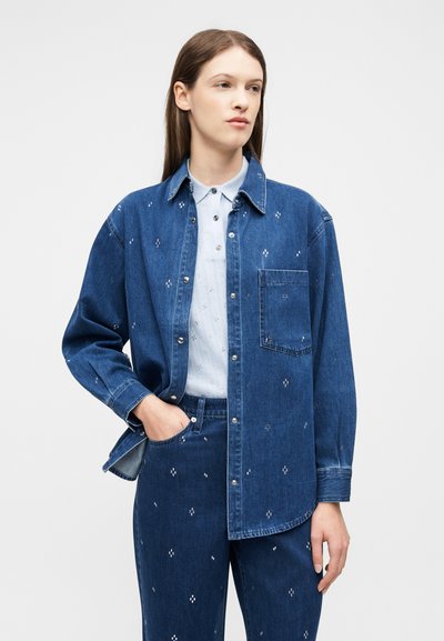 Femme portant une veste en denim bleu foncé assortie à un pantalon de la même couleur avec de petits motifs brodés blancs, et une chemise boutonnée bleu clair.