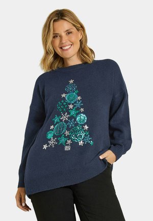 Femme souriante portant un pull bleu marine décoré de formes de sapins de Noël en sequins bleus et argentés et un pantalon noir.