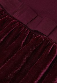 Burgundská zamatová tkanina so zbieraným dizajnom na spodnej časti a hladkým, širokým pásom. Textúra je mäkká a bohatá na farbu.