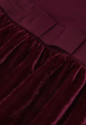 Burgundska baršunasta tkanina s naboranim dizajnom na dnu i glatkim, debljim pojasom. Tekstura je meka i bogata bojom.