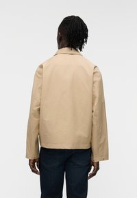 Μ πεζ μπουφάν με καθαρή σιλουέτα, που διαθέτει μυτερό γιακά, μακριά μανίκια και ίσια κοπή. Φορεμένο πάνω από σκούρο μπλε τζιν παντελόνι.