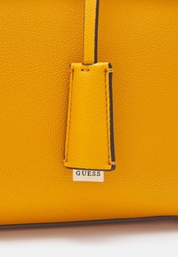 Gros plan d’un sac en cuir jaune texturé avec une bandoulière cousue et une petite plaque métallique portant le nom de la marque Guess.