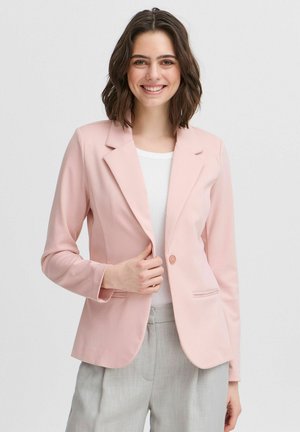 Zachte roze blazer met een enkele knoop, gelapte revers en voorzakken. Draagt over een witte top, gecombineerd met lichtgrijze broek.