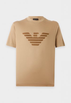 Jeune homme debout portant une chemise à manches courtes beige avec un logo noir à motif, un jean bleu foncé et des baskets noires sur fond blanc.
