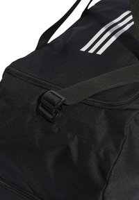Crni torba za putovanje s teksturiranom završnom obradom, s patentnim zatvaračem, plastičnom kopčom i detaljem s tri bijele pruge. Uključena je podesiva traka.