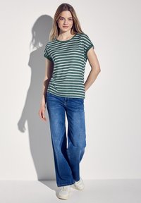 Zielono-biała, w paski, krótka koszulka z długim rękawem w zestawie z szerokimi niebieskimi dżinsami i białymi sneakersami. Posiada podwinięte rękawy i luźny krój.
