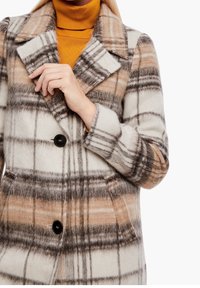 Abrigo a cuadros en colores beige, marrón y crema; hecho de una tela suave con cuello de muesca y detalle de botón negro.