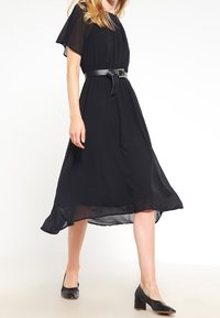Robe midi noire avec des manches en tulle flottantes, taille cintrée par une ceinture et un ourlet fluide. Associée à des chaussures noires à talons blocs.