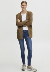Bruine, gestructureerde cardigan over een wit shirt, gecombineerd met blauwe skinny jeans en grijze sneakers. Model staat met gekruiste armen en glimlacht.