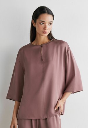 Kvinde med langt mørkt hår iført løstsiddende lilla top med kvartærmer og lille nøglehulshalsudskæring, stående op ad hvid væg.