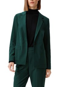 Green blazer made of smooth fabric, featuring a single button closure, peak lapels, and two front pockets. Styled with a black turtleneck underneath.