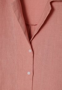 Camicia di lino rosa chiaro con colletto a rotondetto, chiusura a un bottone e tessuto morbido e texture. Dettagli di cucitura visibili sul colletto.