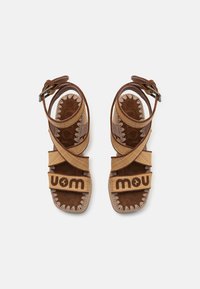 Sandalias de ante marrón y tejido entrelazado con tiras cruzadas, que presentan la palabra "mou" en la parte frontal y costuras decorativas a lo largo de los bordes.