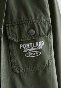 Olivgrüne Arbeitsjacke mit einer Brusttasche, die mit weißen gestickten Text "PORTLAND Workwear" und einem Knopfverschluss versehen ist. Strapazierfähiger Stoff.