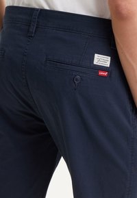 Pantalon Levi's bleu marine foncé avec poche arrière à bouton et étiquettes de marque blanches et rouges sur le côté droit d'une personne portant une chemise blanche.