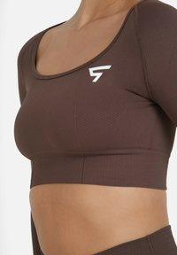Smeđi sportski grudnjak s teksturom, sa izrezom u obliku kupole, prilagođenim dizajnom i bijelim logom na lijevoj strani. Glatka tkanina i podržavajuća struktura.